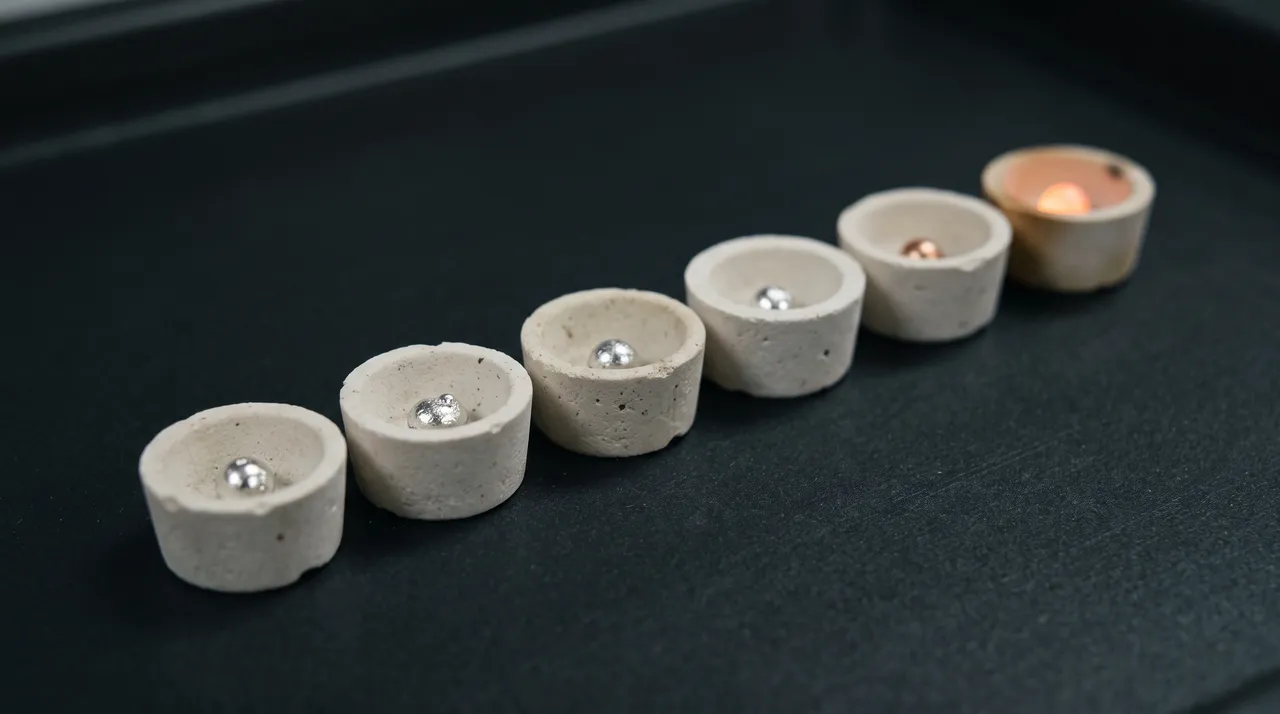 A row of assay cupels on a dark bench, each holding one measured metal bead; the last still glows.